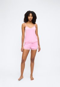 Γυναίκα όρθια, ξυπόλητη, φοράει ροζ καμισόλα με δαντέλα και ταίριαστο σορτς σε απλό λευκό φόντο.