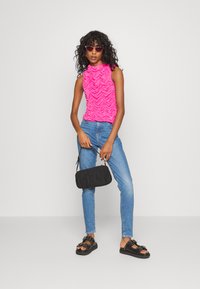 Top sem mangas fuchsia com franzidos texturizados, jeans de denim azul, mala preta e sandálias pretas com tachas decorativas. Cabelo encaracolado e óculos de sol.