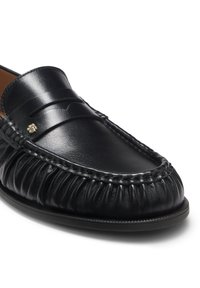 Mocassin penny en cuir noir avec des coutures détaillées le long du bout et un petit logo doré sur la bride.