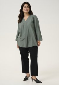 Green, textured blouse with a V-neck, long sleeves, and a relaxed fit, paired with black straight-leg pants and black slingback heels.