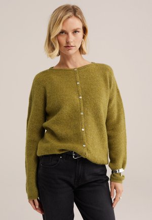 Frau mit kurzen blonden Haaren, die einen grünen, geknöpften Cardigan und schwarze Jeans trägt, steht vor einem schlichten hellen Hintergrund.