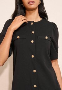 Fekete blúz rövid ujjakkal, kerek nyakkivágással, elöl és a zsebeknél arany virágos gombokkal, amelyek részletgazdagságot és kontrasztot adnak.