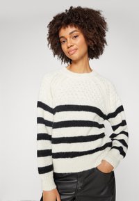 Suéter de punto en crema y rayas negras, con un escote redondo y puños acanalados, hecho de hilo suave y texturizado.