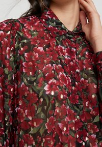 La blusa floral roja transparente presenta un follaje verde, acentos metálicos, un collar clásico y cierres de botones en la parte delantera.