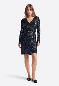 Crna haljina sa šljokicama, dugim rukavima, otvorenim dekolteom i wrap dizajnom, koja ima teksturiranu, sjajnu površinu i završava iznad koljena.