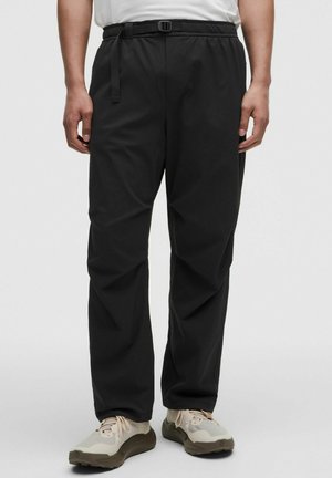 Mann trägt schwarze Freizeithose mit verstellbarem Gürtel und weißen Sneakers vor einfarbigem Hintergrund.