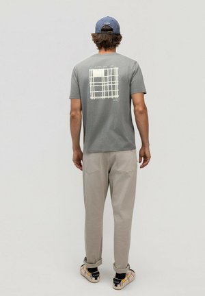 Hombre con camiseta gris con estampado gráfico, pantalones beige, zapatillas de cuadros y gorra azul está de espaldas sobre un fondo liso.