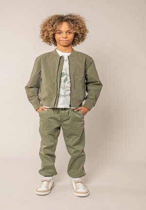 Enfant aux cheveux bouclés portant une veste bomber verte, un pantalon vert, un t-shirt graphique et des baskets blanches, debout les mains dans les poches.