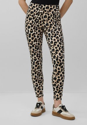 Löwenmuster-Leggings aus tanfarbenem und schwarzem Stoff, mit eng anliegendem Schnitt und elastischem Bund, kombiniert mit beigen Sneakers mit schwarzen Akzenten.