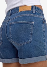 Τζιν σορτσάκια σε σκούρο μπλε με διπλωμένα μανίκια, διαθέτουν δύο πίσω τσέπες και μια μπεζ ετικέτα μάρκας στη μέση. Λείος υφασμάτινος ιστός.