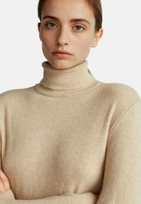 Beżowy sweter z golfem wykonany z miękkiego materiału, o luźnym kroju i minimalnej teksturze. Brak widocznych wzorów czy akcentów.