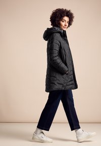 Abrigo acolchado negro con capucha, que presenta paneles cosidos, bolsillos laterales y un corte relajado. Combinado con jeans oscuros y zapatillas blancas.