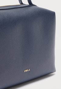 Κοντινό πλάνο μιας γωνίας τσάντας Furla από ναυτικό μπλε δέρμα με υφή, με χρυστό φερμουάρ και μικρό χρυσό λογότυπο σε λευκό φόντο.