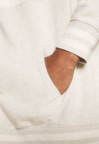 Dunkelhäutige Hand teilweise in der Tasche eines hellbeigen Sweatshirts mit gestreiften Bündchen und Saum.