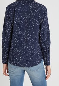 Chemise bleu marine avec un motif de petits points blancs épars, manches longues, poignets à boutons et col classique, associée à un jean bleu clair.