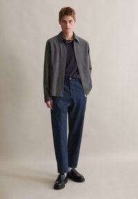 Szara kurtka zapinana na zamek na granatową koszulkę, w połączeniu z luźnymi, ciemnoniebieskimi spodniami dżinsowymi i czarnymi butami, na neutralnym tle.