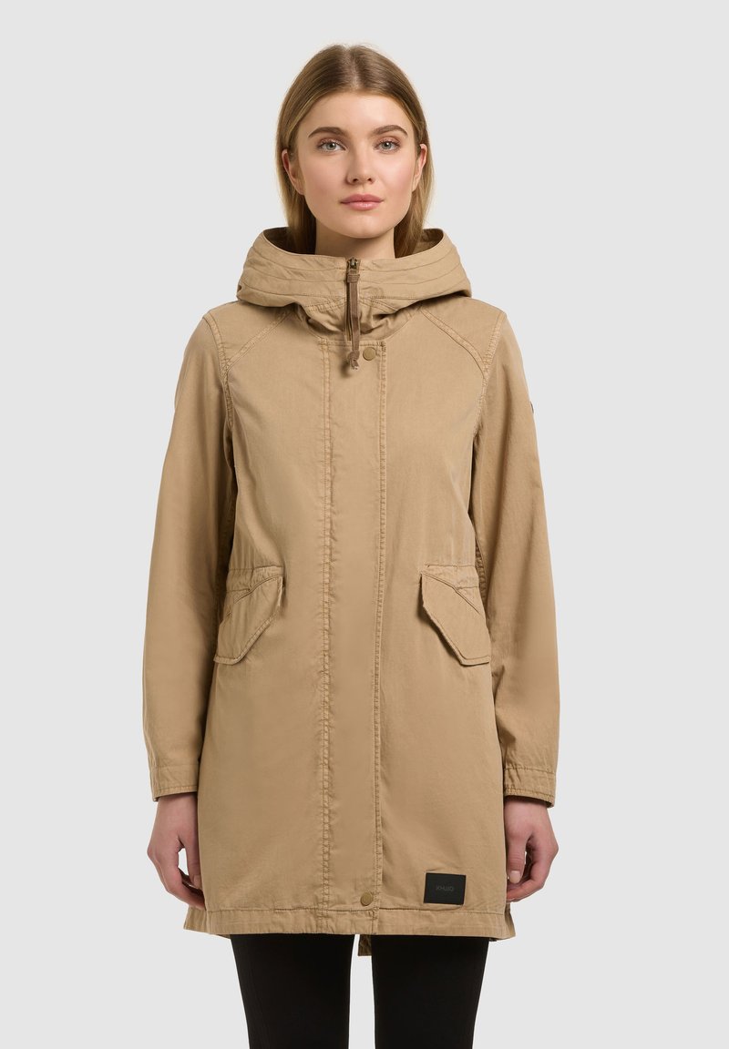 Femme portant un manteau beige à manches longues avec capuche, poches avant et fermeture éclair, debout devant un fond uni.