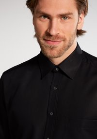 Sort skjorte med knapper og spidse krave, brystlomme og synlige syninger. Glat stof og klassisk pasform.