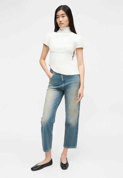 Jeune femme portant un pull à col roulé blanc à manches courtes, un jean bleu délavé et des ballerines noires, debout avec une main dans la poche.