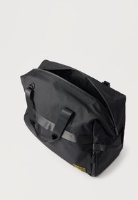 Μαύρη ορθογώνια τσάντα duffel με διπλά χερούλια, ανοιχτό φερμουάρ στην κορυφή, πλαϊνή τσέπη με φερμουάρ και κίτρινο λογότυπο στη γωνία του κάτω μέρους.