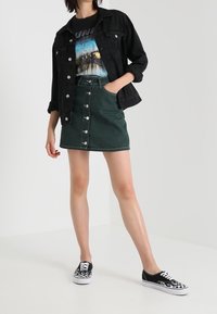 Giacca di jeans nera sopra una maglietta grafica, abbinata a una gonna di jeans verde con bottoni frontali e sneakers a fantasia bianca e nera.