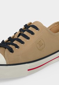 Baskets en toile beige avec lacets noirs, semelle en caoutchouc blanche avec une bande rouge et logo embossé sur le côté.