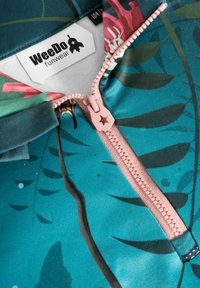 Teal stof med tropiske bladmønstre, der har en pink lynlås og etiket. Glat tekstur med livlige farver og detaljerede designaccentuer.