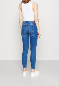 Žena v modrých skinny džínách s vysokým pasem a zadními kapsami, bílé tílko a bílé platformové tenisky, stojící na šedé podlaze.