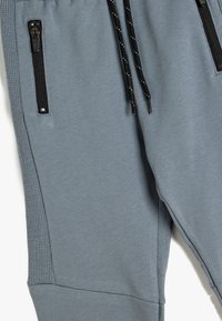 Grijze atletische joggers van zacht materiaal, met zwarte ritsen op de zakken, geribbelde zijpanelen en een verstelbare trekkoord met kleine accenten.