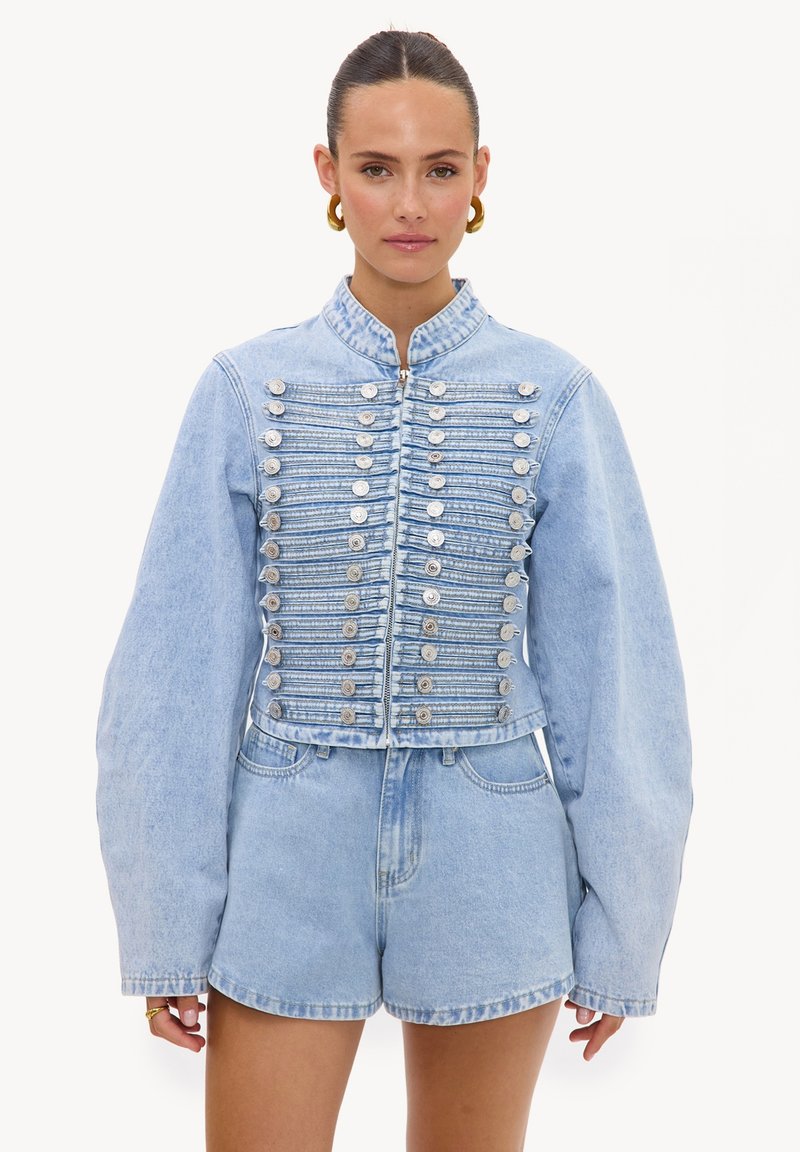 Femme portant une veste en jean bleu clair avec des boutons argentés et un short en jean taille haute assorti, debout sur fond blanc.