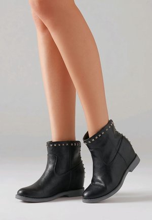 Piernas con botas de cuero negro hasta el tobillo con tachuelas cuadradas plateadas alrededor de la parte superior y trasera, de pie sobre un suelo gris claro.