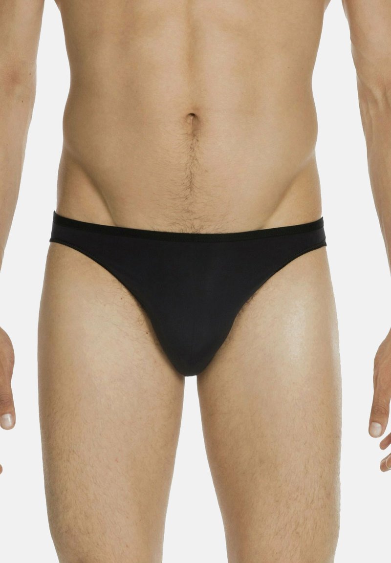 Torso maschile e parte superiore delle gambe con slip neri semplici, in piedi contro uno sfondo chiaro con le braccia rilassate ai lati.