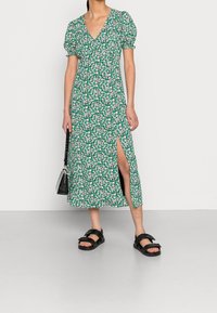 Zelené květinové šaty s nabíranými rukávy, hlubokým výstřihem do V a rozparkem až ke stehnům, v kombinaci s černými sandály a malou kabelkou.