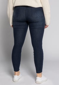Jeans ajustados de un azul oscuro con cintura alta, dos bolsillos traseros y sutiles detalles de costura en la parte delantera. Llevados con zapatillas blancas.