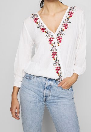 Mujer con blusa blanca de cuello en V con bordado floral rojo y púrpura combinada con vaqueros de tiro alto azul claro, mano en el bolsillo.