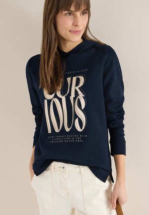 Femme portant un sweat à capuche bleu marine avec le texte « Curious » et un pantalon blanc, debout avec une main dans la poche contre un fond uni.