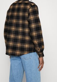 Camicia di flanella con motivo a quadri nero e beige, dotata di colletto, polsini con bottoni e vestibilità rilassata. Indossata con jeans azzurri chiari.