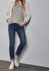 Ljus beige bomberjacka över en salviagrön topp, kombinerad med mörkblå slim fit-jeans och vita sneakers. Jeanssen är uppvikta vid anklarna.