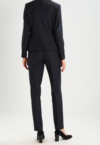 Marinblå kostym med en figursydd blazer och rak byxa. Tillverkad av textilt material, med eleganta linjer och subtila sömmar, kombinerad med svarta klackar.