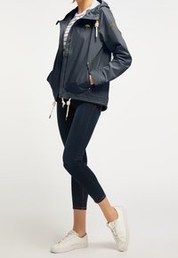 Marinblå vattentät jacka med huva, två blixtlåsfickor, kontrasterande guldaccents och flätade dragsnören, kombinerad med mörka jeans och vita sneakers.