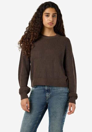 Jeune femme aux longs cheveux bouclés portant un pull en tricot marron et un jean bleu sur un fond clair uni.