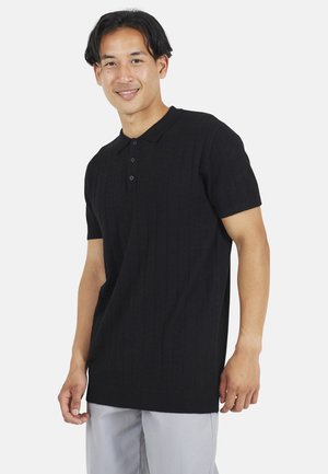 Schwarzes Polo-Shirt mit kurzen Ärmeln und gerippter Textur, Drei-Knopf-Leiste und entspannter Passform, kombiniert mit hellgrauen Hosen.
