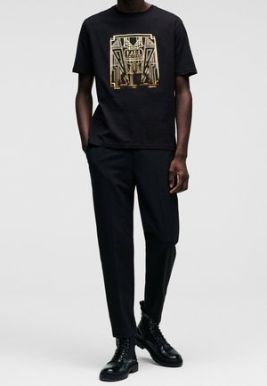 Mężczyzna w czarnym t-shircie z grafiką i złotym nadrukiem, czarnych spodniach i czarnych sznurowanych butach wojskowych stoi z rękami w kieszeniach.