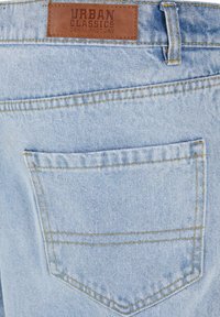 Svijetloplave traperice s pravokutnom kožnom oznakom na kojoj piše "Urban Classics Denim Factory" iznad stražnjeg džepa. Vidljive detalje šivanja.