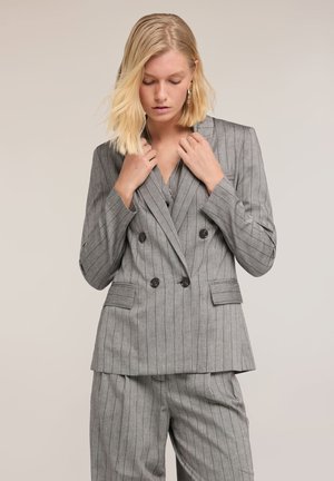 Blondine vrouw die de kraag aanpast van een grijze dubbelgeknoopte blazer met krijtstrepen en bijpassende broek tegen een neutrale achtergrond.