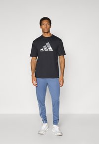 T-shirt noir à manches courtes avec un logo Adidas réfléchissant, associé à un pantalon de sport bleu avec des rayures blanches sur les côtés. Des baskets blanches complètent le look.