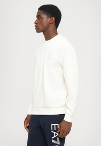 Sudadera de color crema con cuello redondo, puños y dobladillo acanalados. Presenta un pequeño logotipo en relieve en el pecho. Textura suave.
