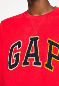 Persona che indossa una felpa rossa brillante con grandi lettere blu navy e gialle "GAP" ricamate sul petto.