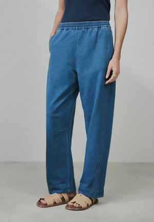 Person wearing loose blue pants with elastic waistband and beige open-toe sandals, standing on a plain light floor and wall background.