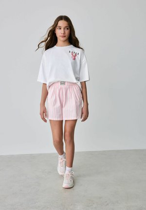Mädchen, das ein weißes übergroßes T-Shirt mit Blumenmuster und rosa-weiß gestreifte Shorts mit elastischem Bund sowie Turnschuhe trägt und läuft.